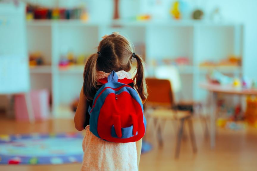 A young girl with pigtails viewed from behind, wearing a red and blue backpack while standing in a bright kindergarten classroom.
