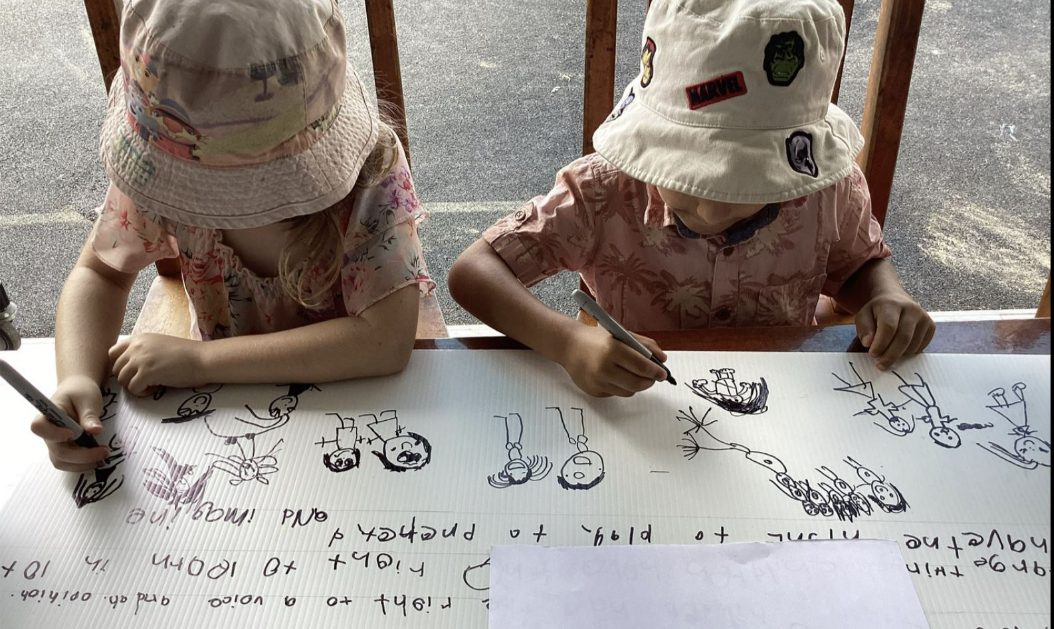 Two young children wearing hats drawing stick figures on a large paper to create the Children's Charter of Rights