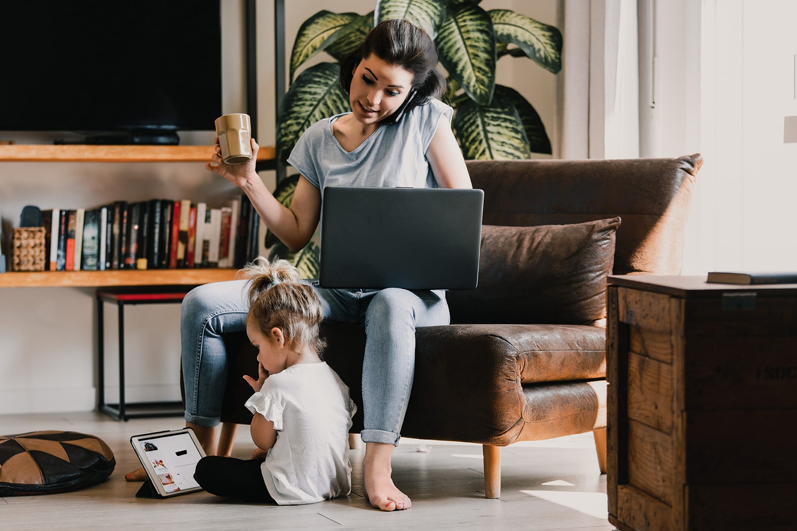 A woman sitting on a leather chair, working on a laptop, holding a mug, and talking on a smartphone, while a young child sits on the floor in front of her looking at a tablet