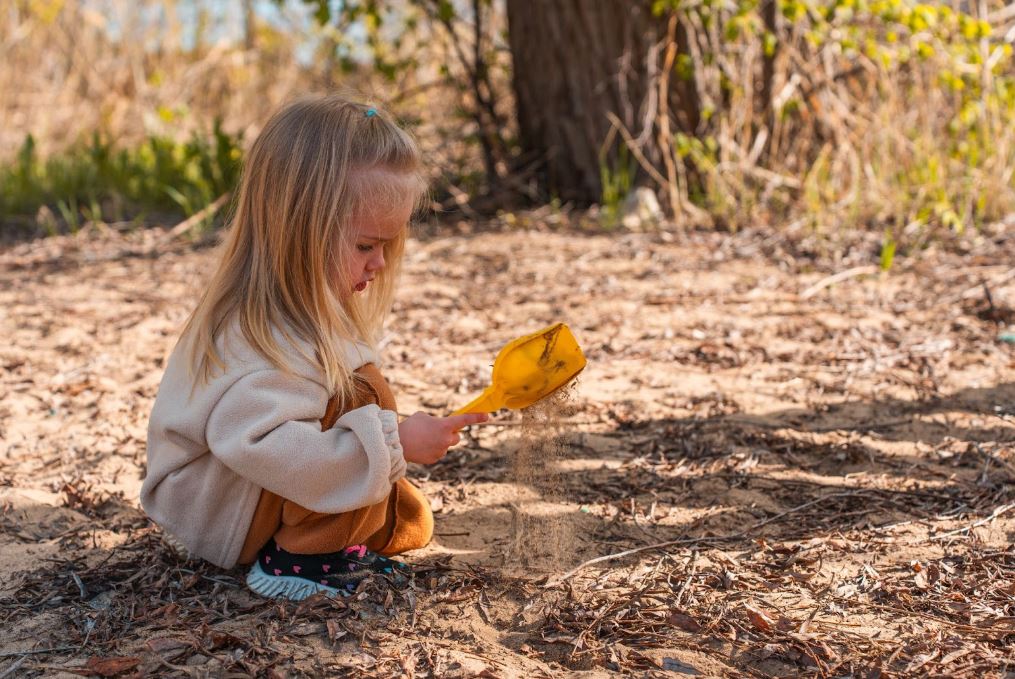 A young girl squatting on the ground digging in the dirt with a yellow toy shovel.