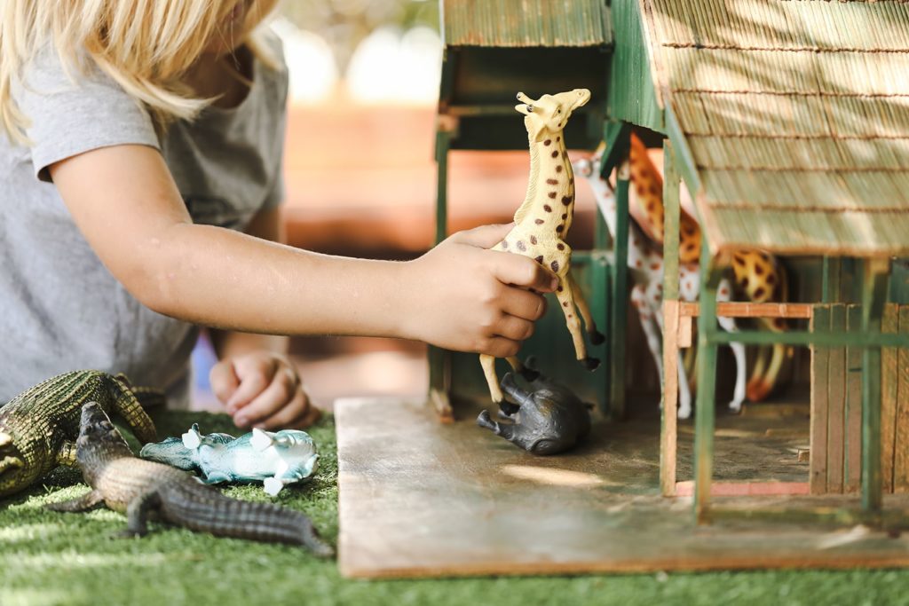 Child playing with a toy giraffe and toy crocodiles near a small wooden stable.